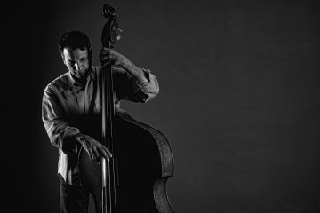 Black-and-white photo of a man playing a double bass, eyes closed, wearing a light shirt, focused on the instrument.