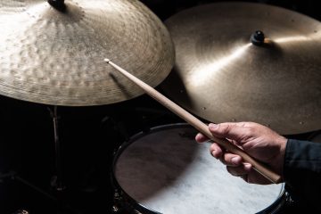 Close-up of a drummer's hand gripping a wooden drumstick above a snare drum with cymbals in the background