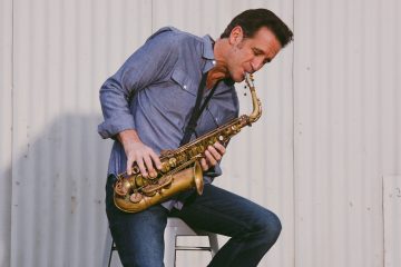 Man in a blue denim shirt and jeans sits on a stool, playing a brass saxophone against a light metal wall.