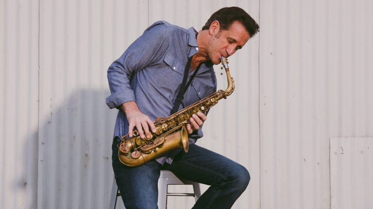 Man in a blue denim shirt and jeans sits on a stool, playing a brass saxophone against a light metal wall.