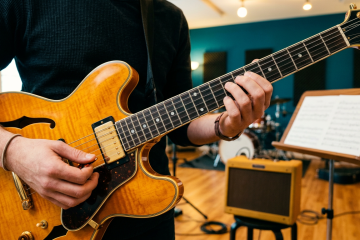 Musician playing an amber semi-hollow guitar in a rehearsal room with sheet music on a stand in the background.