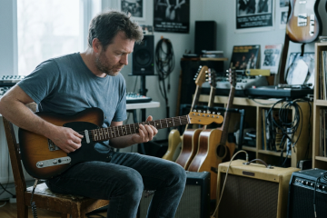 Man in a blue t-shirt sits on a wooden chair, playing a sunburst electric guitar in a home studio with amps, guitars, records visible.