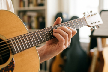 Close-up of a person’s fretting hand on the strings of an acoustic guitar, in a cozy music room.