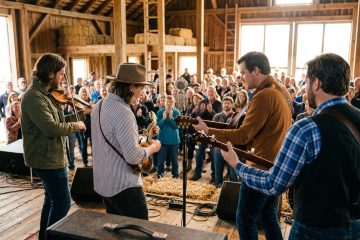 Four musicians perform on a wooden stage in a rustic barn, playing string instruments as a crowd applauds.