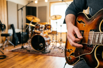 Close-up of a guitarist playing a sunburst semi-hollow guitar in a rehearsal studio, with drums in the background.