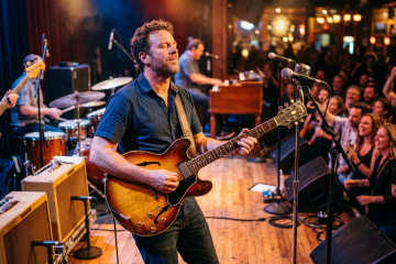 Guitarist in a blue shirt playing a sunburst semi-hollow guitar on stage as a lively crowd watches from the right.
