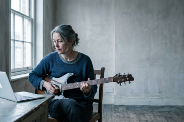 Guitarist studying a Keith Wyatt lesson on her laptop in a sparse practice room, practicing the mute technique