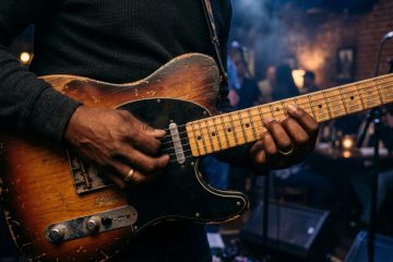 Close-up of a blues guitarist's hands playing a boogie shuffle on an electric guitar on a dimly lit stage, worn finish visible on the body.