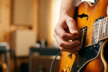 Close-up of a guitarist's hand tightening a string on a sunburst electric guitar, with an amp blurred in the background.
