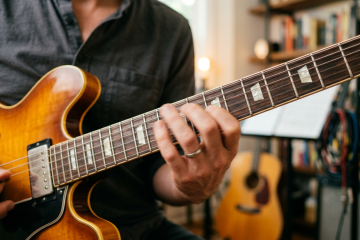 Close-up of a person playing a sunburst electric guitar, fretting chords on a wooden neck in a cozy studio setting.