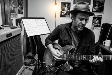 Guitarist caught mid-strum in a dimly lit practice room, pick angled upward on a return stroke over a semi-hollow electric guitar body