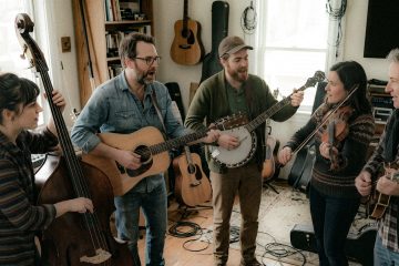 Five musicians jam in a cozy living room, playing guitar, banjo, violin, and upright bass together.