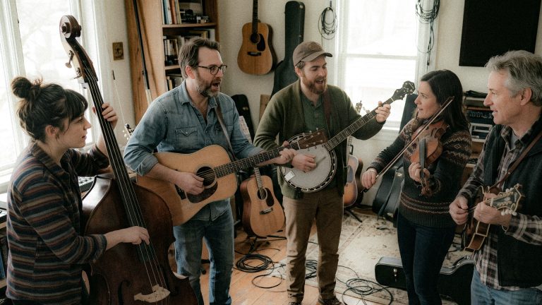 Five musicians jam in a cozy living room, playing guitar, banjo, violin, and upright bass together.
