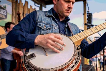 Close-up of a man in a denim jacket plucking a banjo on an outdoor stage, with another guitarist blurred in the background.