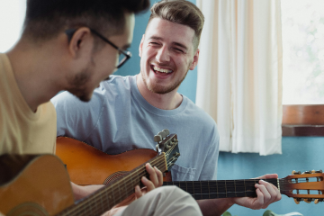 Two men playing acoustic guitars and laughing together in a sunlit room by a window.