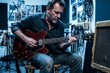 a blues guitarist in his 50s sitting slightly off-center on a wooden stool in a small rehearsal room, hunched forward mid-strum on a semi-hollow electric guitar, a battered combo amp clipped at the right edge of the frame