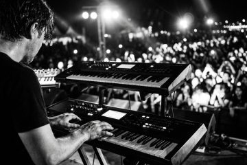 Black and white photo of a keyboardist on stage playing a pop progression to adoring fans at a live festival.