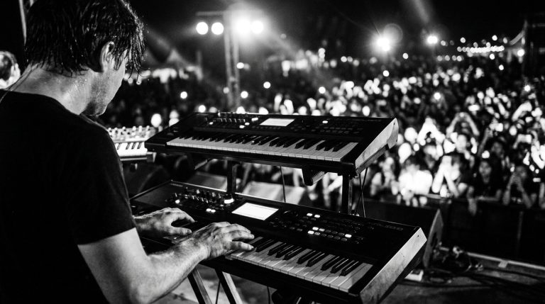 Black and white photo of a keyboardist on stage playing a pop progression to adoring fans at a live festival.