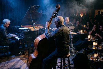 on stage shot of a jazz trio performing at a small cocktail bar, upright bass player, pianist and drummer playing the changes.