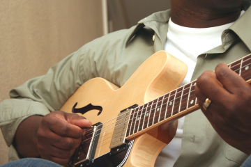 Close-up of a person playing a light wood electric guitar, focusing on the hands on the strings and fretboard.
