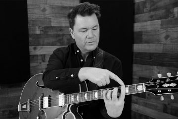 Black and white photo of Sean McGowan as he demonstrates essential technique for fingerstyle jazz guitar in a teaching studio, pointing to his left hand finger position