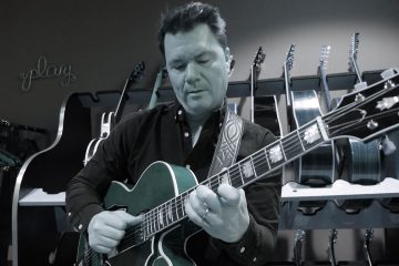 Sean McGowan demonstrates shadowing with the thumb, tinted desaturated photo of Sean playing guitar with a rack of guitars behind him in a greenroom