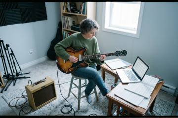 Woman in her 50s sits in a sparsely decorated practice room playing a guitar plugged into a small practice amp, laptop open, learning fingerstyle guitar