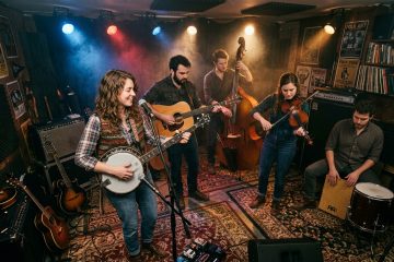Folk band performing in a cozy studio: singer with banjo at center, guitar, upright bass, violin, and percussion players around her under colorful stage lights.