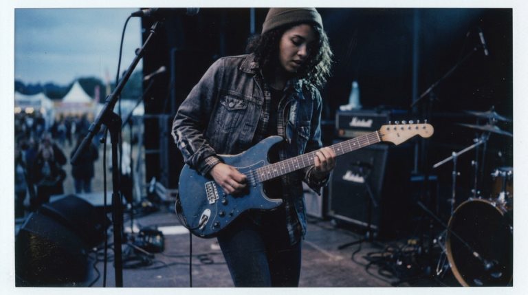 a young female guitar player solos on an outdoor stage demonstrating triplet fills over a blues shuffle groove