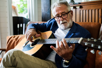 Older man with glasses plays an acoustic guitar on a brown sofa in a bright living room.