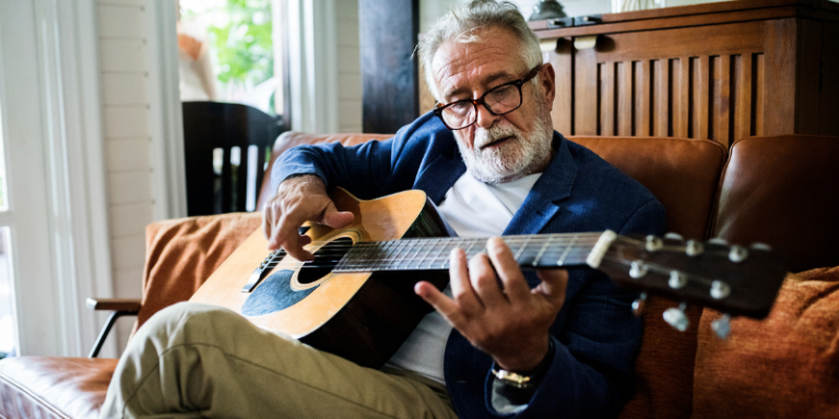 Older man with glasses plays an acoustic guitar on a brown sofa in a bright living room.