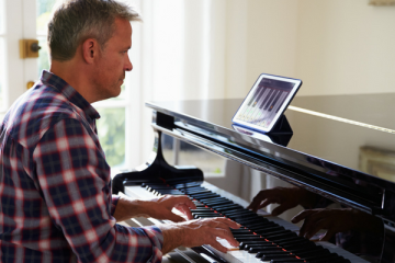 Side view of a man in a plaid shirt playing a glossy black grand piano with a tablet displaying music on the lid.