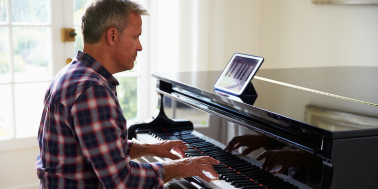 Side view of a man in a plaid shirt playing a glossy black grand piano with a tablet displaying music on the lid.