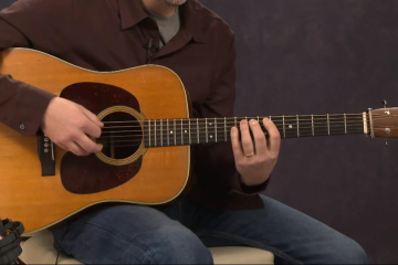 Person playing an acoustic guitar, with fingers on the fretboard while seated on a chair.