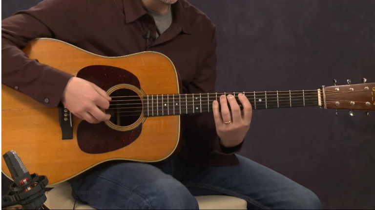 Person playing an acoustic guitar, with fingers on the fretboard while seated on a chair.