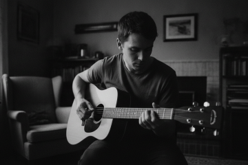 black and white photo of a young man playing acoustic guitar in his bedroom, looking down at his left hand - to illustrate a common neck posture