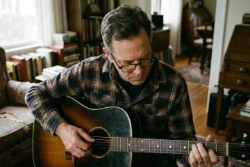 A man in his 50s practices his upstroke technique on acoustic guitar in his home office